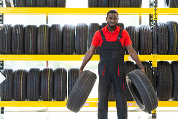 Black male Mechanic holding a tire and showing wheel tires at car repair service and auto store shop. Expertise mechanic working in automobile repair garage.