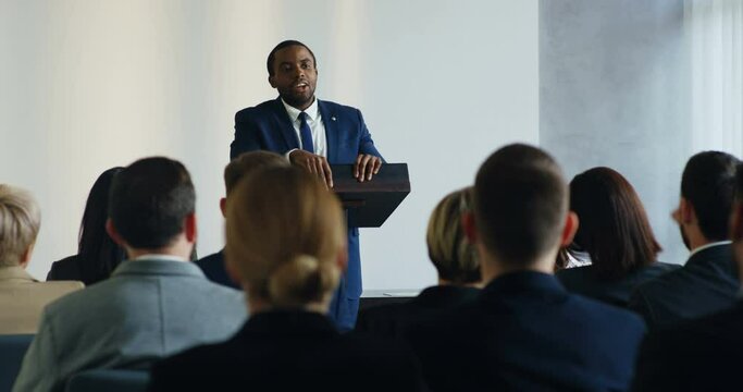 Male African American business speaker standing at the lecturn at the conference and interacting with people in the hall during speech while someone from the audience rising hand and asking the