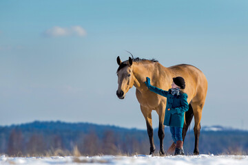 Beautiful girl standing near her Trakehner yellow horse outside in field. Child with a big mare standing together on snow in winter background