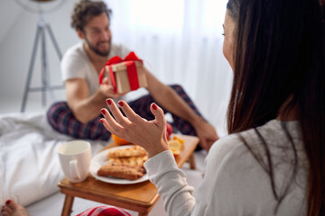 A young guy giving a Xmas present to his excited girlfriend sitting in the bed. Christmas, relationship, love, together