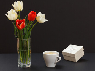 Bouquet of yellow tulips in vase with gift box and cup on a black background.