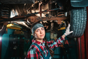 Fototapeta na wymiar Portrait of a young female mechanic in uniform and gloves, posing near a car that is under repair. In the background is an auto repair shop