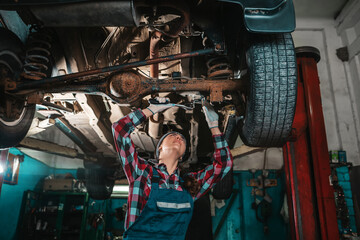 Obraz premium Portrait of a young female mechanic in uniform and gloves, repairing a car. Bottom view. The car is located on the lift