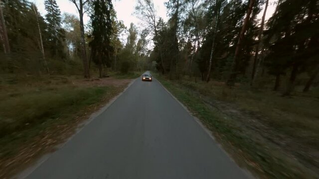 Black jdm car with headlights driving on asphalt road coniferous forest, aerial cinematic shooting fpv drone. Traveling vehicle moving on countryside way in pine woodland in summer evening twilight.