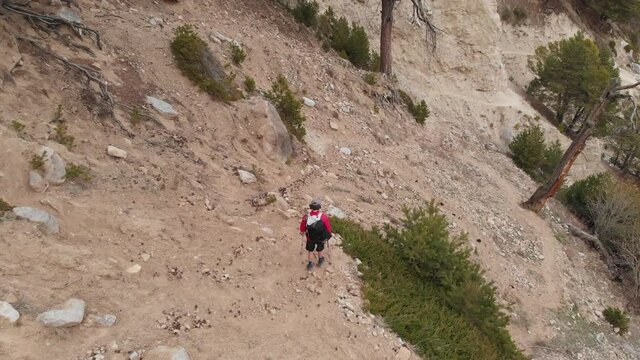 Aerial view of a man in a cap and shorts and with a backpack walks with trekking poles along a mountain valley high in the mountains