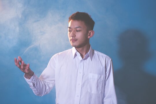 Handsome Thoughtful Young Man Gesturing By Smoke Against Blue Background