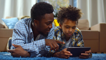 African american man and son at home lying on floor in living room and using tablet pc