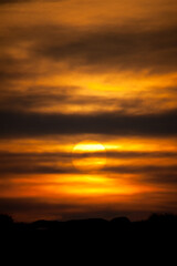 A setting sun behind layers of cloud and deep orange sky tones with a black horizon.Vertical Image