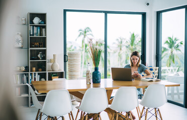 Young female freelancer messaging on smartphone while using laptop at home
