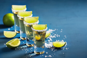 Shots with traditional Mexican golden tequila, decorated with salt and lime, stand on a blue concrete surface, copy space, horizontal orientation.