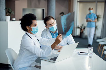 Fototapeta premium Black female doctor and her colleague analyzing X-ray while wearing face masks at medical clinic.