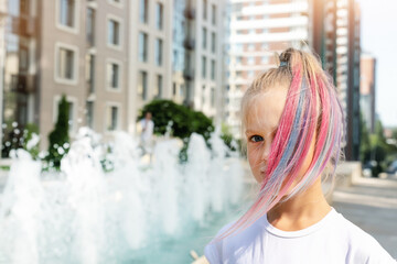 Cute adorable caucasian little kid girl with multicolored pastel chalk pained dyed blond hair on city street with fountain background. Female child portrait pink colorful hairstyle smiling outdoors