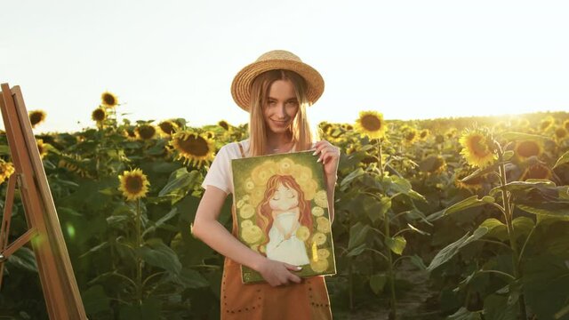 The woman is holding the picture and smiling. There is an easel next to it. She stands in the middle of a field of sunflowers. 4K