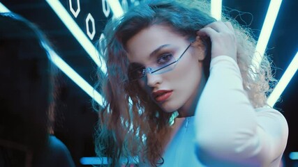 Portrait beautiful trendy woman wear white outfit and futuristic glasses posing looking into camera standing by illuminated wall in night club. Cyberpunk style, sensual look clubber in neon blue light