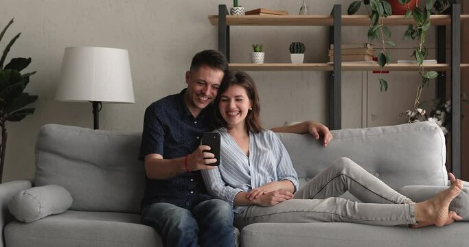 Millennial romantic couple relax on couch at home, happy husband showing on smartphone to wife funny videos, take selfie family picture, make video call having fun using wireless modern tech concept