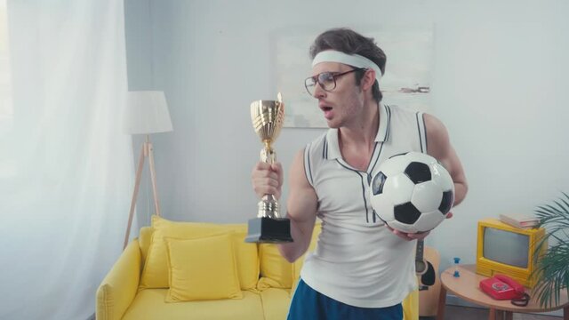 Excited footballer with trophy cup and soccer ball, celebrating win at home