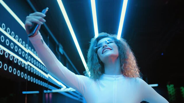 Portrait beautiful trendy woman wear white outfit and futuristic glasses posing take selfie photo using smartphone standing by illuminated wall in night club. Cyberpunk style, clubber in neon light