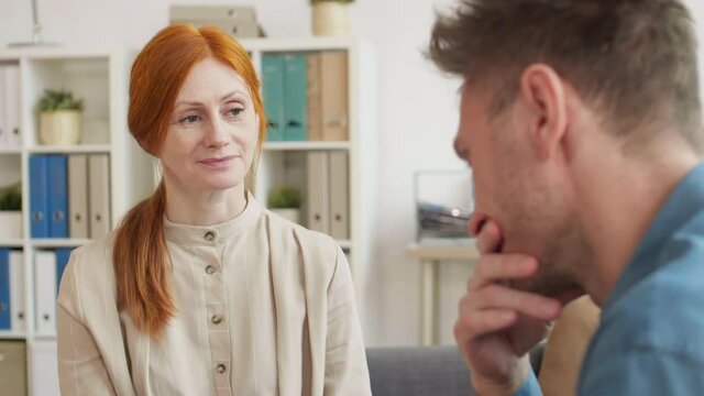 Medium close-up of red-haired Caucasian woman conducting psychological therapy to cropped man sitting in front of her in office, people talking