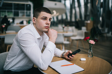 Young pensive businessman with mobile and notepad in cafe