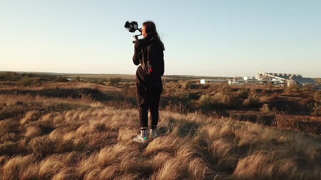 Shooting a girl with camera on stab who filming wind farms on sunset. Modern styled content creator on a hills. A spirit of freedom in art.