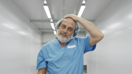 Portrait of happy aged surgeon removing safety mask and cap walking in hospital corridor - Powered by Adobe