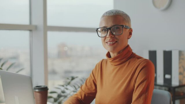Chest up shot of attractive mid-aged businesswoman with short grey hair using laptop and posing for camera with smile while working in office