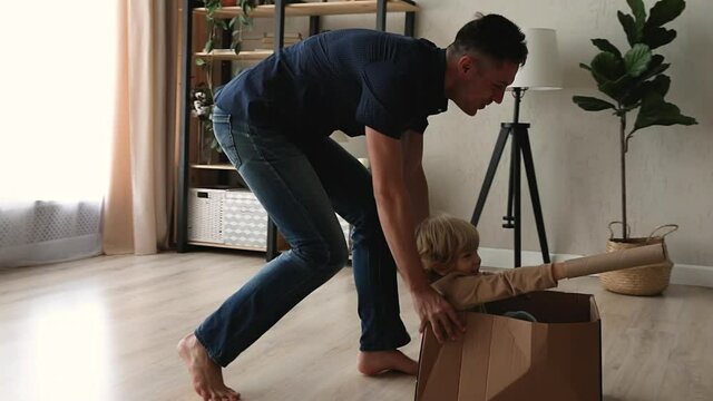 Father ride 4s preschool son while he sitting inside of big cardboard box, happy lively family laughing play active game at modern warm living room, celebrate relocation day to new comfy home concept