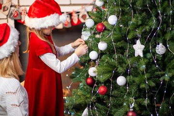 a girl in a red dress and a santa claus hat near the fireplace clings a toy on the christmas tree