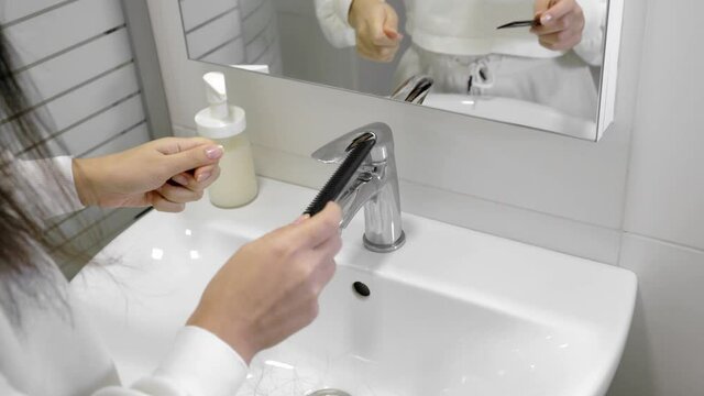 Excessive hair shedding when combing. The girl combs her hair in front of the bathroom mirror. A lot of hair remains on the comb and in the sink
