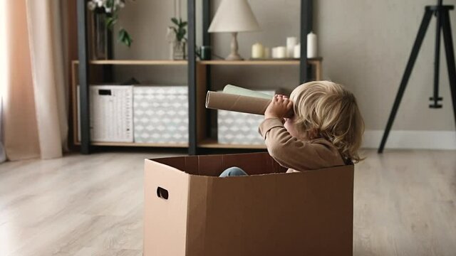Cute little kid boy sit inside of cardboard box holding paper tube looking into distance imagines himself discoverer. Preschool curious child holding carton handmade binoculars play game alone at home