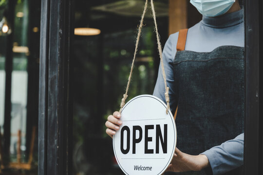 Restart. friendly waitress woman wearing protection face mask turning open sign board on glass door in modern coffee shop, cafe restaurant, retail store, small business owner, food and drink concept