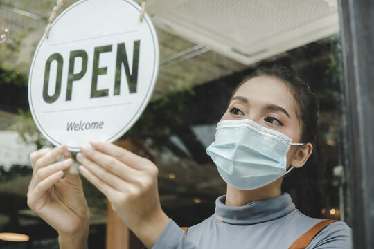 Reopen. friendly waitress wearing protection face mask turning open sign board on glass door in modern cafe coffee shop, cafe restaurant, retail store, small business owner, food and drink concept