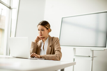 Young woman working on laptop in bright office with big screen behind her