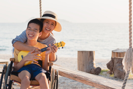 Asian special child on wheelchair is singing, playing ukulele happily on the beach with parent,Natural sea beach background,Life in the education age of disabled children,Happy disability kid concept.