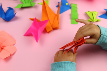 A child makes origami from colored paper on a pink background