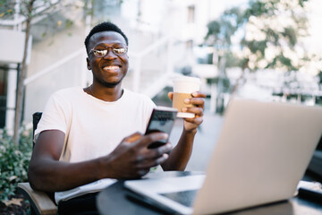 Smiling african american hipster guy in eyewear enjoying remote job on coffee terrace holding smartphone and coffee cup, positive dark skinned male freelancer sending text messages on cellular