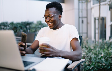 Happy dark skinned hipster guy millennial holding mobile phone reading news and information from social networks, cheerful african american man enjoying 4G connection for watching video on smartphone