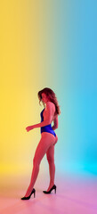 Beautiful seductive girl in fashionable blue swimsuit on gradient yellow-blue background in neon light. Full-length portrait. Copyspace for ad. Summer, fashion, beauty, emotions concept. Flyer