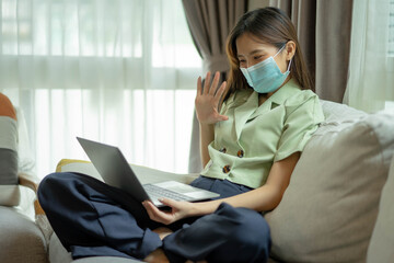Young woman in medical mask sitting on the sofa in her own house is video call conference with...