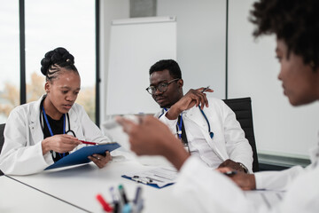 Team of three African-American doctors sitting in modern hospital and talking during business meeting.