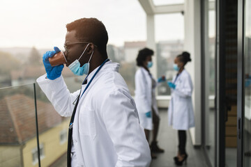 Three Afro American doctors standing on hospital balcony during coffee break or work time out. They wearing protective face masks as virus pandemic protection. - Powered by Adobe