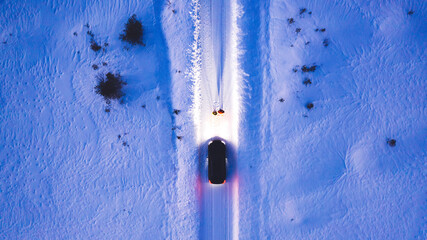 Aerial top view of romantic couple of tourists standing near car while headlights lighting them way on in winter darkness, bird's eye view of vehicle in snowy north lands. Wanderlust in road trip