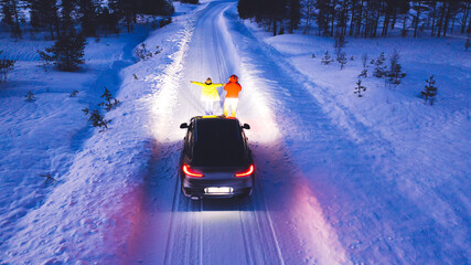 Aerial back view of romantic couple of tourists standing near car while headlights lighting them way on in winter darkness, bird's eye view of vehicle in snowy north lands. Wanderlust in road trip
