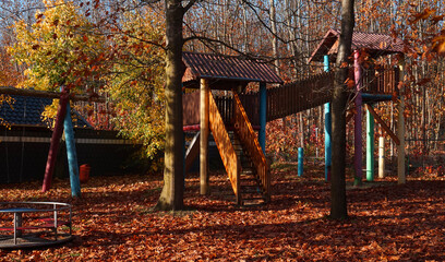 Abandoned colourful children's playground in the fall. A swing, carousel and climbing house are visible. Combined with giant American oaks that have lost their leaves. It is slowly getting cold.
