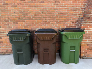 3 trash bins sit on cement driveway beside the house in front of brown bricks wall as background....