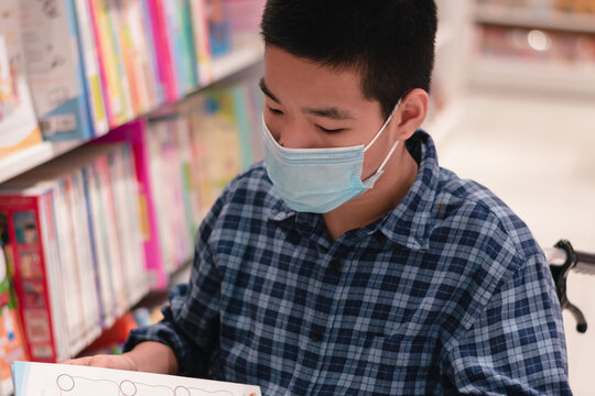 Asian Disabled Child On Wheelchair Wearing A Mask Reading A Book From Shelves In Books Store Or School Library, Lifestyle In The Education Age Of Special Need Kids, Happy Disability Kid Concept.