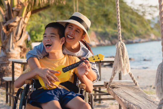 Asian special child on wheelchair is singing, playing ukulele happily on the beach with parent,Natural sea beach background,Life in the education age of disabled children,Happy disability kid concept.