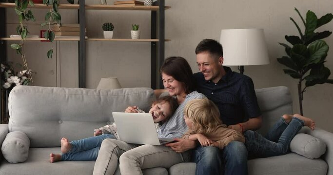 Relaxed couple their preschool son and daughter rest on comfy sofa in living room spend weekend laughing watching family movie or cartoons on laptop, enjoy free time at home, modern tech usage concept