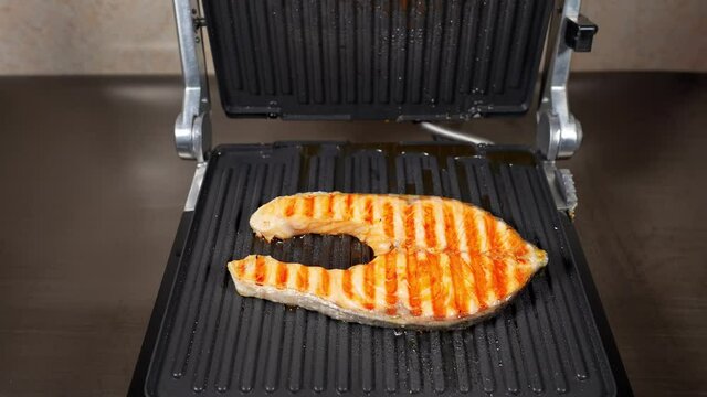 The grill lid opens. A piece of salmon fish is cooking on the electric grill