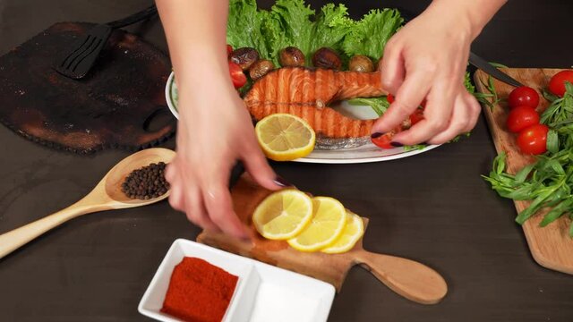 Woman's hand puts slised  lemons on the plate with fried salmon, lettuce, potatoes and halfed cherry tomatoes.